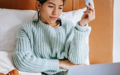 Une jeune femme se détend au lit, fait des achats en ligne avec un ordinateur portable et une carte de crédit en main.