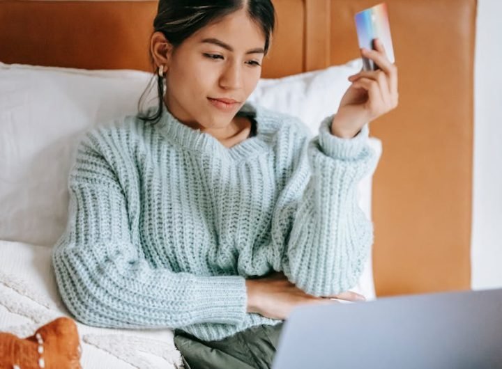 Une jeune femme se détend au lit, fait des achats en ligne avec un ordinateur portable et une carte de crédit en main.